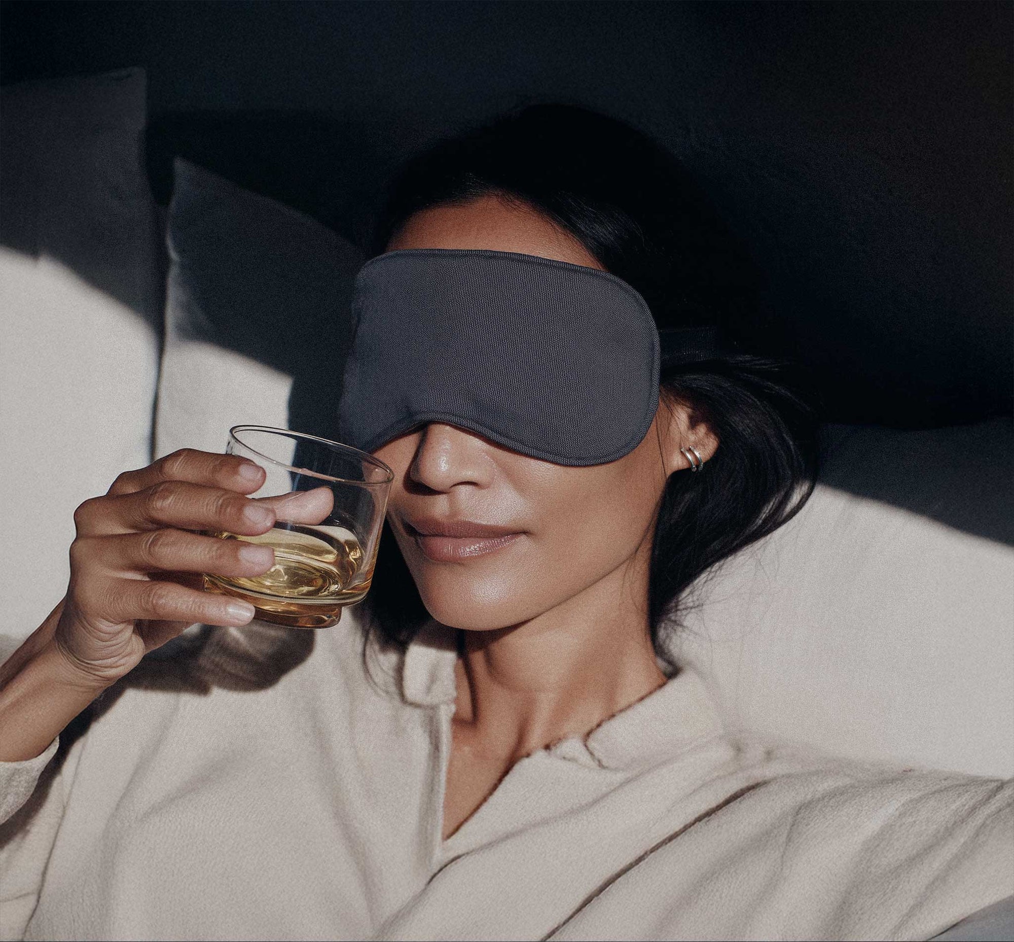 Person wearing a sleep mask and holding a glass of liquid in a cozy setting.